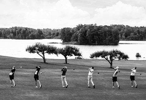 Fem golfspelare står på rad på en golfbana vid sjön. 